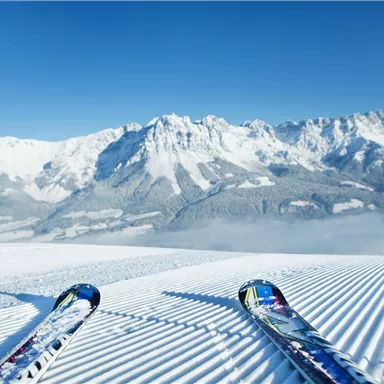Eine atemberaubende Winterlandschaft mit schneebedeckten Bergen im Hintergrund. Vorne sind Skier auf der perfekt präparierten Piste zu sehen.