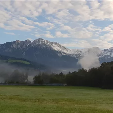 A picturesque mountain landscape with snow-capped peaks and gentle clouds. In the foreground, a green meadow stretches.
