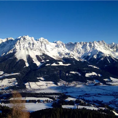 Eine beeindruckende Berglandschaft mit schneebedeckten Gipfeln und klarem blauen Himmel. Im Vordergrund sind grüne Täler zu sehen.