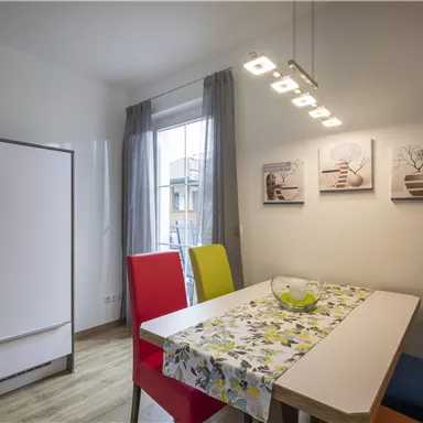 Eine moderne Küche mit einem Esstisch und bunten Stühlen. An der Wand hängen drei Bilder, und das Licht ist hell und einladend.