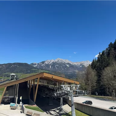 Eine moderne Gondelbahn mit einer markanten Holzarchitektur. Im Hintergrund sind majestätische Berge und ein klarer blauer Himmel zu sehen.