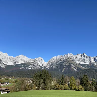 An impressive mountain landscape with snow-capped peaks and a clear blue sky. In the foreground, green meadows and a few houses can be seen.