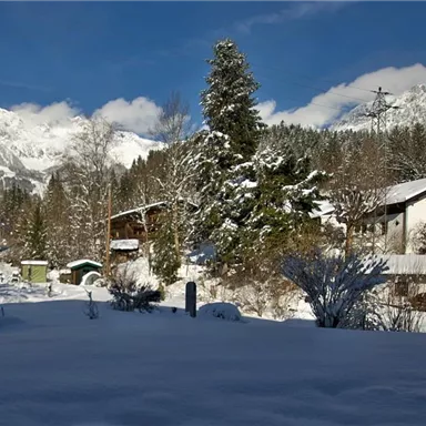 A winter landscape with snow-covered houses and beautiful mountains in the background. The sky is clear and blue.