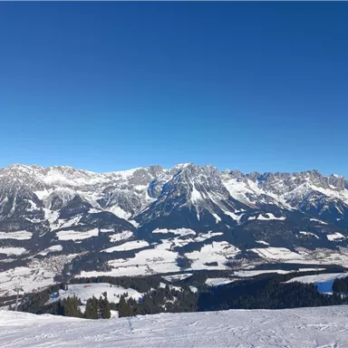 Eine winterliche Berglandschaft mit schneebedeckten Gipfeln und klarem, blauem Himmel. Im Vordergrund liegt eine schneebedeckte Ebene.