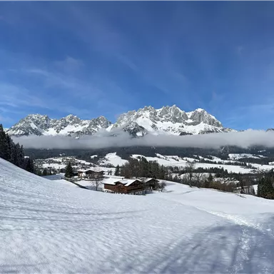 A winter landscape with snow-covered hills and impressive mountains in the background. The sky is clear and blue, creating a calm atmosphere.
