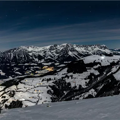 A snow-covered mountain landscape under the night sky. In the distance, the lights of a small village shine.