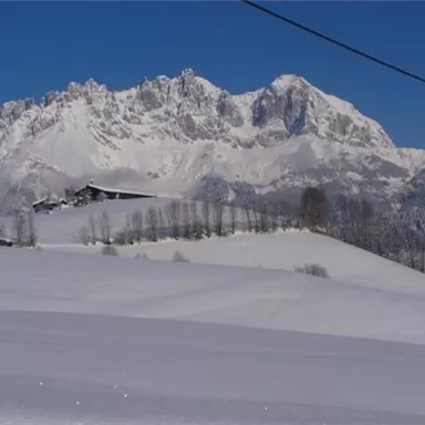 A white winter landscape with snow-covered hills. In the background, an impressive mountain range rises under a clear blue sky.