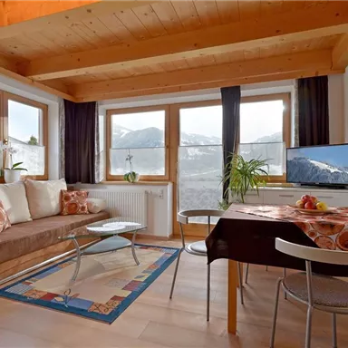 Cozy living room with wooden ceiling and large windows offering a view of the mountains. There is a sofa, a dining table, and a television in the room.