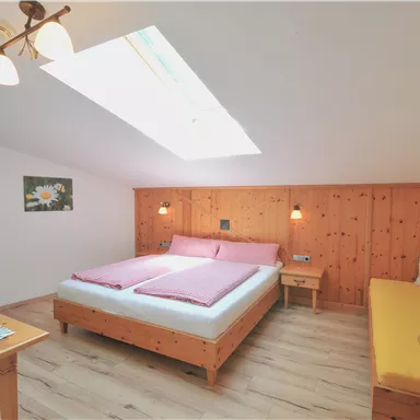 Ein gemütliches Zimmer mit einem Doppelbett und einer Sitzecke. Die Wände sind aus Holz und ein Dachfenster sorgt für viel Tageslicht.