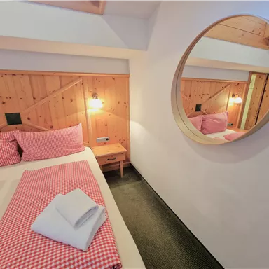 Ein gemütliches Schlafzimmer mit einem Doppelbett und karierten Bettwäsche in Rot. Der runde Spiegel und die Holzwände schaffen eine warme Atmosphäre.