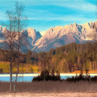 A picturesque landscape with mountains in the background and a clear lake in the foreground. Deciduous trees and meadows complete the picture.