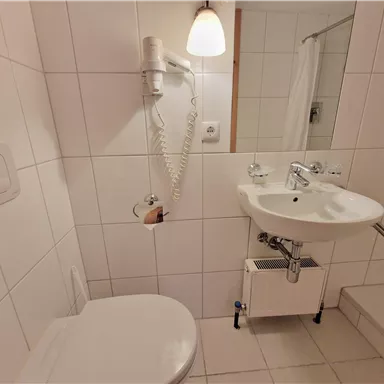 Ein einfaches Badezimmer mit weißen Fliesen. Es verfügt über eine Toilette, ein Waschbecken und eine Dusche.