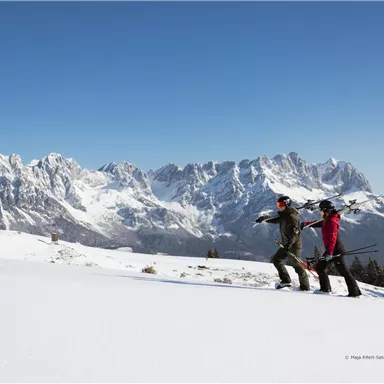 Two skiers are standing on a snow-covered area, enjoying the view of the majestic mountains. The sky is clear and blue.