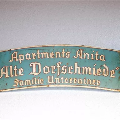 A decorative sign with the inscription "Apartments Anita 'Old Village Smithy' Family Unterrainer." The sign is designed in a rustic style.