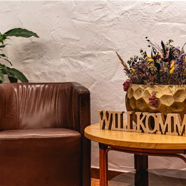 Eine gemütliche Ecke mit einem braunen Stuhl und einem Tisch. Auf dem Tisch steht eine dekorative Vase mit Trockenblumen und dem Schriftzug "WILLKOMMEN".