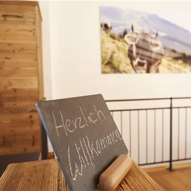 Ein Willkommensschild aus schwarzem Holz steht auf einem Tisch. Im Hintergrund ist ein Bild einer Kuh auf einer Wiese zu sehen.