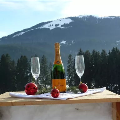 A bottle of sparkling wine is on a wooden table in the snow, surrounded by two glasses and red Christmas ornaments. In the background, snow-covered mountains and forests can be seen.