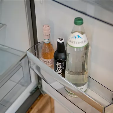 Ein Kühlschrankinnenteil mit drei Getränken: eine Flasche Roséwein, ein schwarzes Getränk und ein Flasche Mineralwasser. Die Regale sind leer und ordentlich.