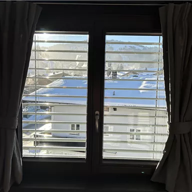 Ein Fenster mit Lamellen, das den Blick auf ein schneebedecktes Dach und die umliegenden Berge freigibt. Die Sonne scheint durch das Fenster und beleuchtet den Raum.