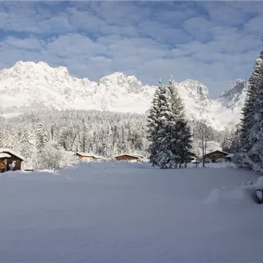 Eine verschneite Landschaft mit hohen Bergen im Hintergrund. Gemütliche Holzhütten sind zwischen den Tannen versteckt.