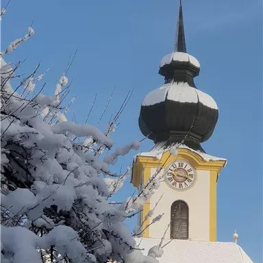 A church with a distinctive spire, covered in snow. The sky is blue and clear.