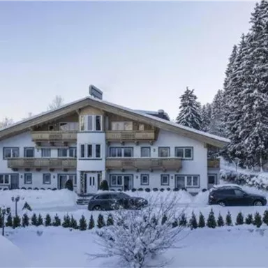 Ein charmantes Chalet im Winter, umgeben von schneebedeckten Bäumen. Vor dem Gebäude stehen zwei Autos.