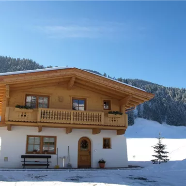 A cozy wooden house in the snow with a large balcony. In the background, forested mountains are visible under a clear blue sky.