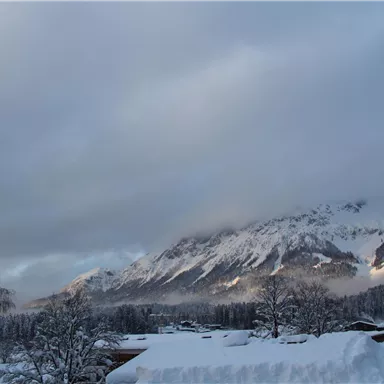 Eine winterliche Landschaft mit schneebedeckten Bergen und tief hängenden Wolken. Der Schnee liegt ruhig auf den Dächern und Bäumen.