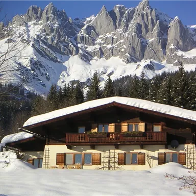 Ein charmantes Haus im Schnee mit einem schneebedeckten Dach. Im Hintergrund erheben sich majestätische Berge.