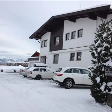 Ein modernes Gebäude im Schnee mit mehreren Autos auf der Auffahrt. Im Hintergrund sind Berge und bewölkter Himmel sichtbar.