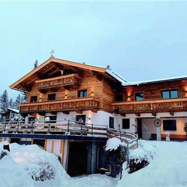 A nice wooden house in the snow, surrounded by fir trees. The facade is cozy and inviting.