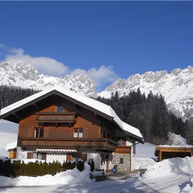 Ein charmantes Holzhaus im Schnee, umgeben von majestätischen Bergen. Der klare blaue Himmel lässt die winterliche Landschaft strahlen.