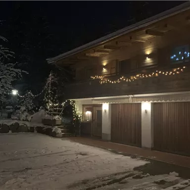 Ein gemütliches Haus im Schnee, beleuchtet von warmen Lichtern. Die Umgebung ist winterlich und einladend.