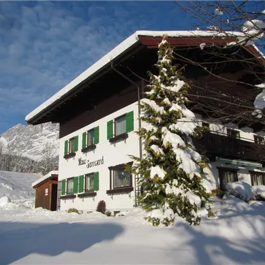 A charming house covered in snow in winter. The surroundings showcase mountains and clear, blue skies.