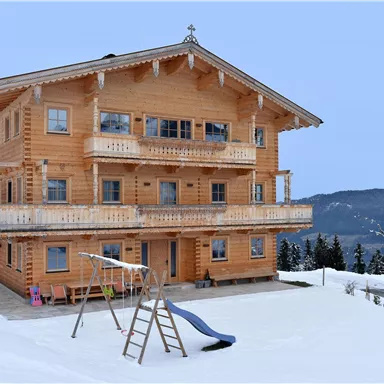 A cozy wooden house in the snow, surrounded by mountains. In front of the house, there is a slide and a swing for children.