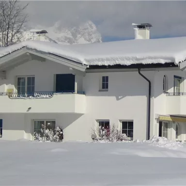 Ein gemütliches Haus im Schnee, umgeben von einer winterlichen Landschaft. Der Himmel ist klar und sonnig, was einen schönen Kontrast zum weißen Schnee bildet.