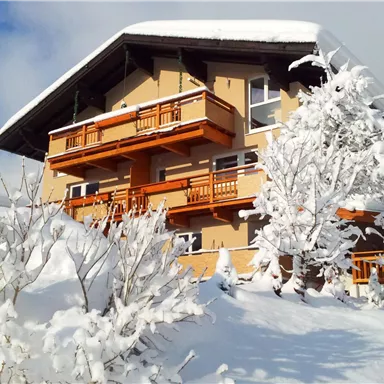 A charming house in the snow with wooden decorations and balconies. The surroundings are characterized by white snow and snow-covered trees.