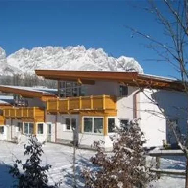 Ein modernes Gebäude im Schnee mit großen Fenstern und Balkonen. Im Hintergrund sind schneebedeckte Berge zu sehen.