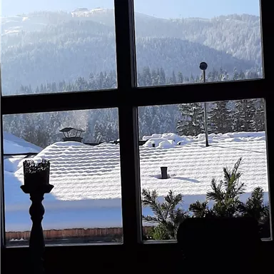 Ein Blick aus einem Fenster auf schneebedeckte Dächer und verschneite Berge im Hintergrund. Der Himmel ist klar und blau.