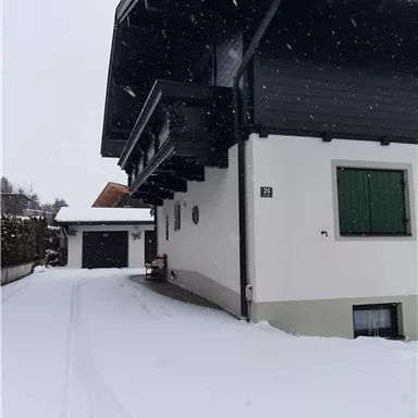 Ein modernes Haus in einer schneebedeckten Landschaft. Die Szenerie ist ruhig und winterlich, mit schneebedeckten Wegen und sanften Schneeflocken.