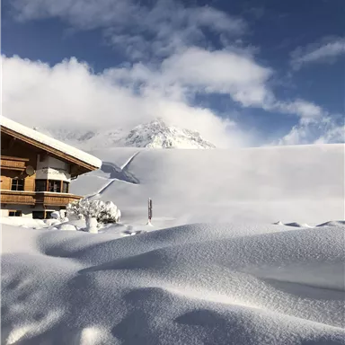 Eine malerische winterliche Landschaft mit einem Holzhaus im Vordergrund. Im Hintergrund sind schneebedeckte Berge und ein blauer Himmel zu sehen.
