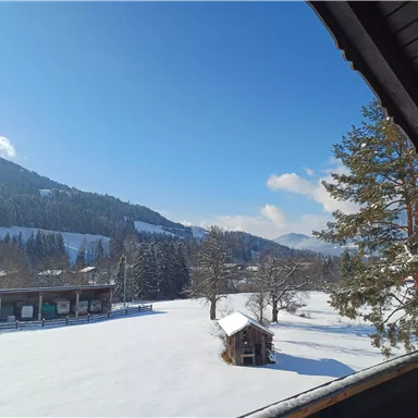 A snowy landscape with mountains in the background and a blue sky. In the foreground, there is a small wooden shed and some trees.