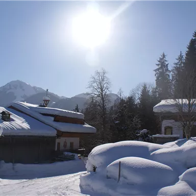 Eine winterliche Landschaft mit schneebedeckten Dächern und sonnigem Himmel. Im Hintergrund sind Berge und hohe Bäume zu sehen.