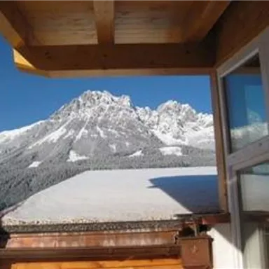 Ein beeindruckendes Bergpanorama mit schneebedeckten Gipfeln und klarem, blauem Himmel. Im Vordergrund ist ein Holzbalcony sichtbar.