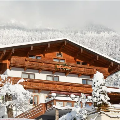 Ein charmantes, holzverkleidetes Chalet im Schnee. Im Hintergrund sind schneebedeckte Berge und ein klarer Himmel zu sehen.