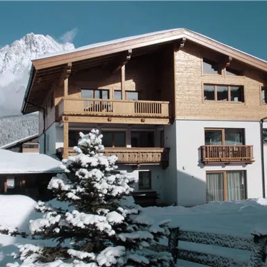 Ein schönes Holzhaus im Schnee mit Balkonen und einem schneebedeckten Garten. Im Hintergrund sind die Berge sichtbar.