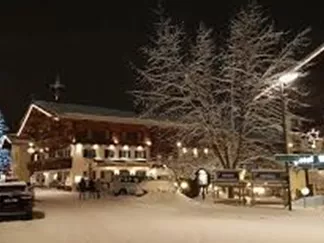 Eine winterliche Szenerie mit schneebedeckten Straßen und festlich beleuchteten Gebäuden. Im Hintergrund steht ein beleuchteter Baum, der eine gemütliche Atmosphäre schafft.