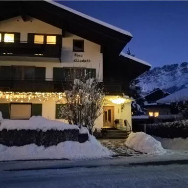 Ein gemütliches Haus mit Weihnachtslichtern, umgeben von Schnee. Im Hintergrund sind majestätische Berge zu sehen.