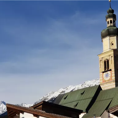 A historic church tower with a distinctive dome in front of an impressive mountain landscape. The sky is clear and blue, creating a peaceful atmosphere.