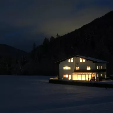 Ein beleuchtetes Haus steht in einer verschneiten Landschaft bei Nacht. Im Hintergrund sind Berge und ein klarer Himmel zu sehen.